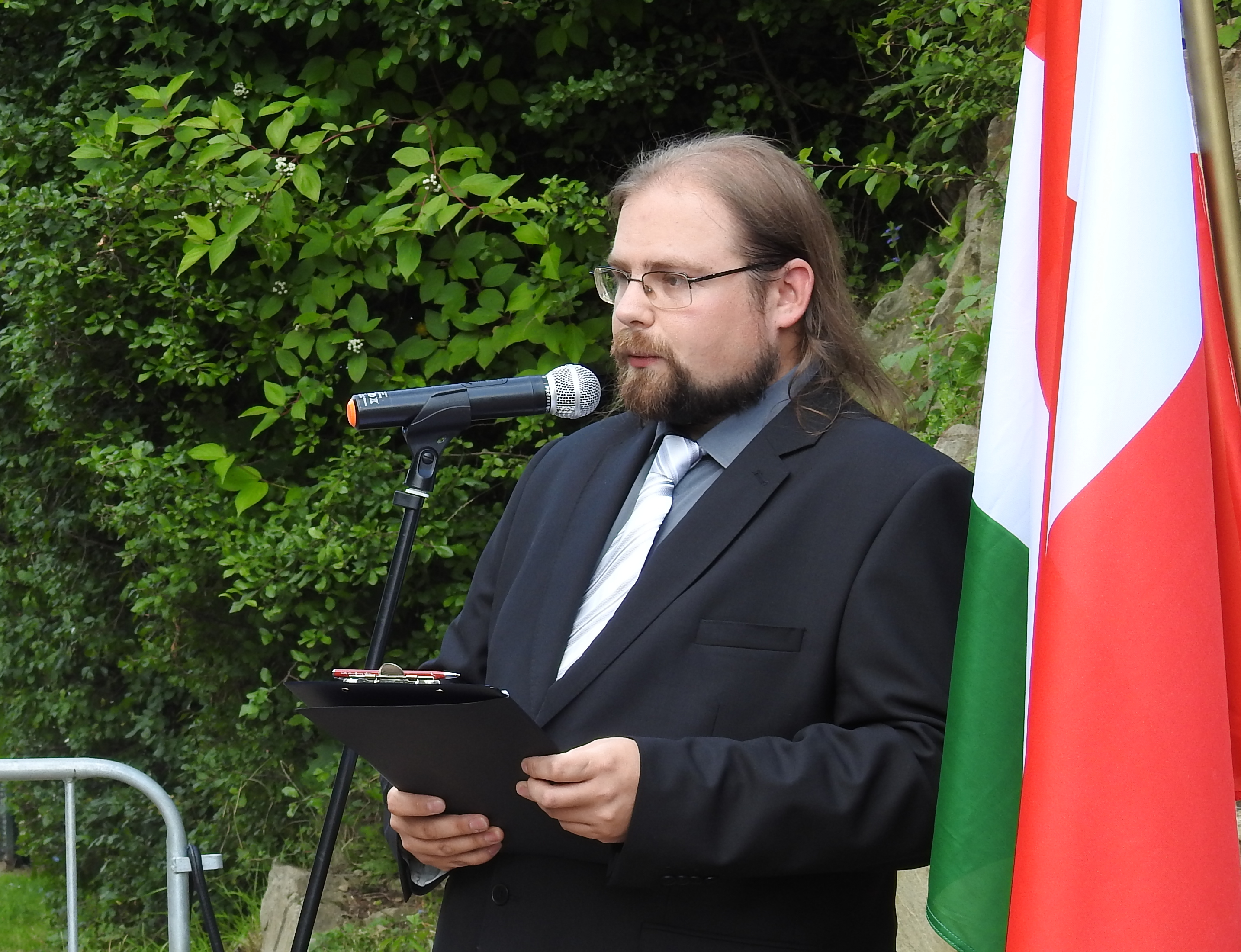 Zdjęcie przedstawia mężczyznę w okularach i czarnym garniturze, mówiącego do mikrofonu, w tle kolorowe flagi i drzewa.