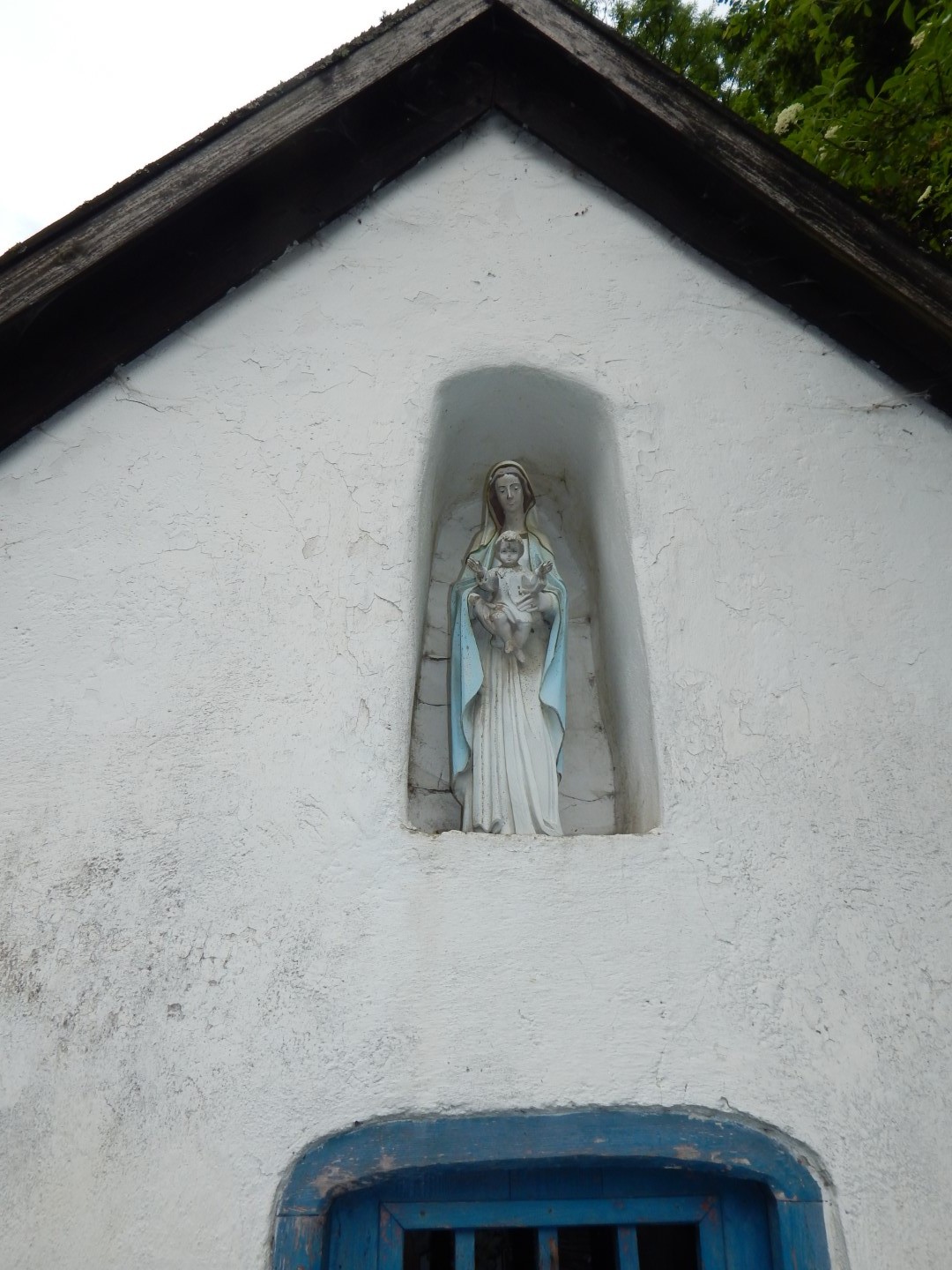Zdjęcie przedstawia figurę Matki Boskiej z Dzieciątkiem w białej wnęce. Wyżej widać szczyt dachu, a poniżej część niebieskich drzwi. 