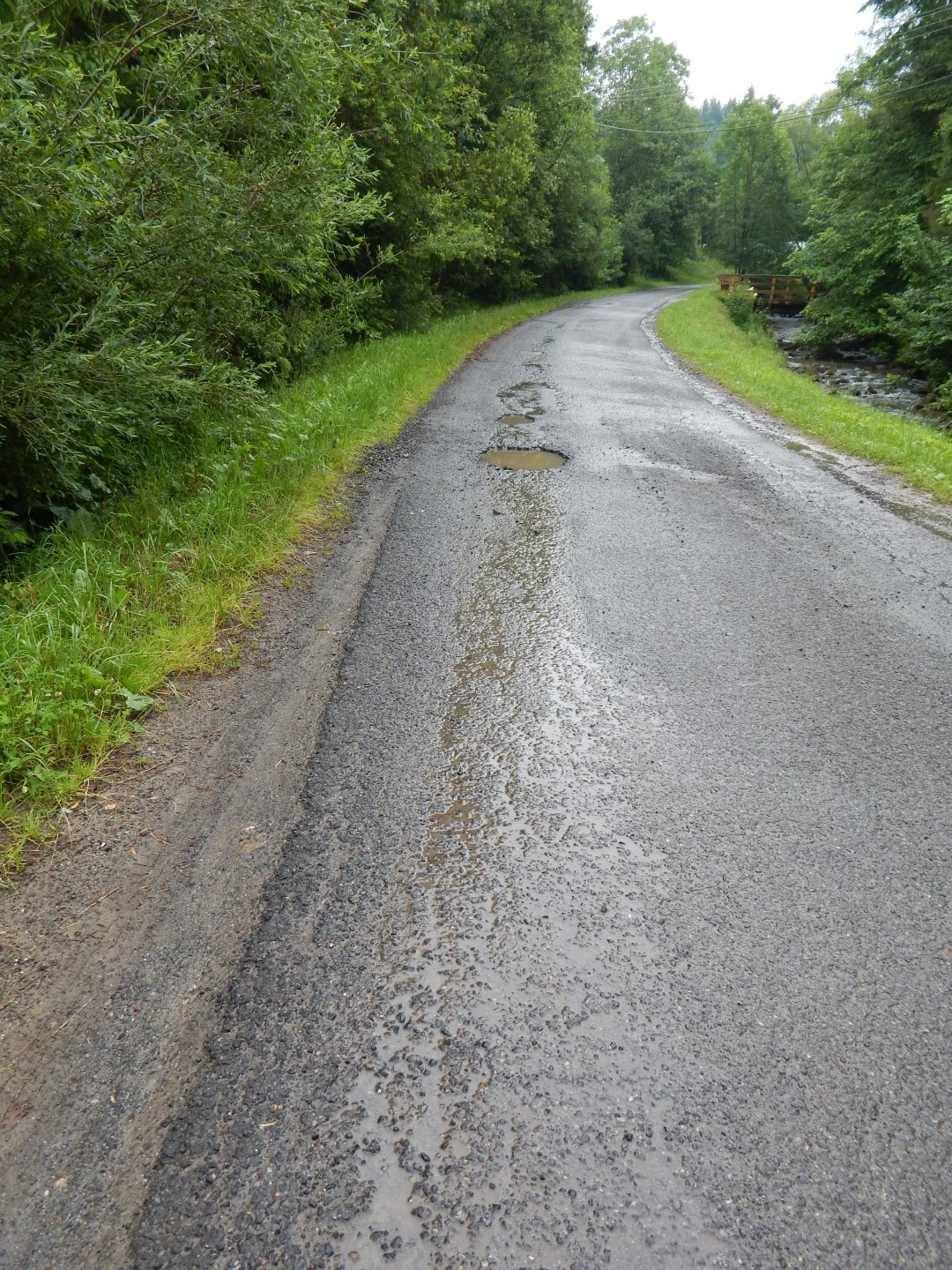 Na zdjęciu znajduje się asfaltowa droga z dziurami wypełnionymi wodą w lewej części drogi. Po prawej stronie płynie strumyk i widać drewniany most w oddali. Wokół drogi rośnie trawa i drzewa.